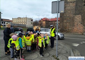 dzieci ubrane w kamizeli odblaskowe stoją przy przejściu dla pieszych wraz z komisarzem lwem i policjantką