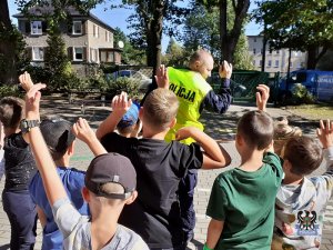 dzieci wspólnie z policjantem uczą się prawidłowego rozglądania przed przejściem przez drogę