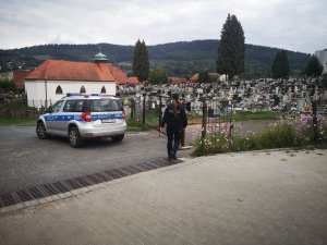 Na zdjęciu widać oznakowany radiowóz policyjny i policjantkę, która idzie w stronę cmentarza znajdującego się w tle