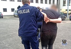 Na zdjęciu widać policjanta prowadzącego zatrzymanego, który ma kajdanki założone na ręce trzymane z tyłu