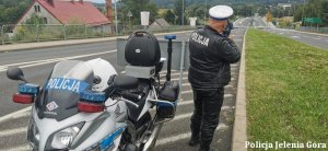 Policjant dokonuje pomiaru prędkości stojąc obok policyjnego motocykla