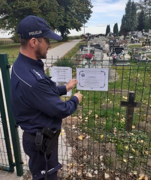 Policjant przyczepia klepsydrę na ogrodzeniu cmentarza