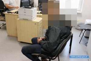 zatrzymany mężczyzna w kajdankach zapiętych z przodu, ubrany w ciemne spodnie i ciemną kurtkę siedzący na krześle, w tle meble biurowe, monitory,  z prawej strony biały napis na niebieskim tle lublin.policja.gov.pl