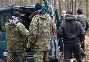 dwóch mężczyzn w umundurowaniu moro stoi przy tylnych drzwiach busa. Obok pojazdu stoją mężczyźni odwróceni plecami