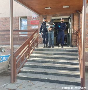 Umundurowani policjanci prowadzą zatrzymanego mężczyznę z kajdankami po schodach przed jednostką Policji.