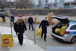 Wrocławscy policjanci noszą paczki dla dzieci w czapkach mikołaja.