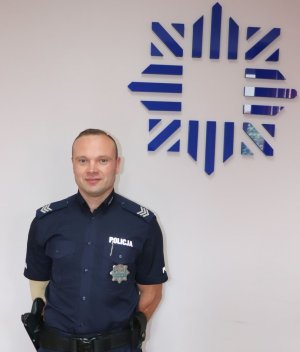 policjant umundurowany stoi na tle ściany na której widnieje granatowy emblemat policji