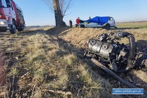 na pierwszym planie silnik pojazdu leżący w przydrożnym rowie po lewej samochód straży pożarnej w oddali przykryty niebieską plandeką uszkodzony pojazd koloru srebrnego, poza drogą