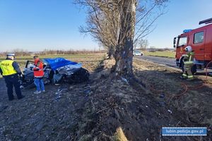 pojazd poza drogą ze zdeformowaną karoserią przykryty niebieską plandeką, policjant w kamizelce wykonujący czynności oraz ratownik medyczny stojący przy samochodzie, po prawej na drodze samochód straży pożarnej