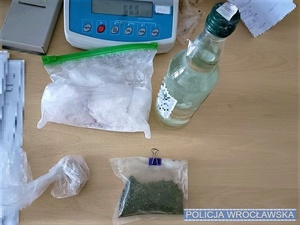 Na stole leżą: woreczek z białym proszkiem, woreczek z zielonym suszem, butelka z przeźroczystym płynem i waga elektroniczna