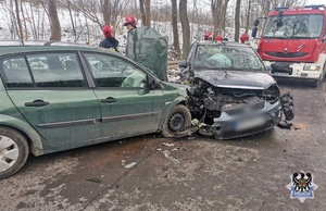 Na zdjęciu widzimy miejsce wypadku drogowego, dwa uszkodzone pojazdy stoją wbite w siebie. W tle stoją strażacy