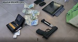 na podłodze leżą broń i pieniądze zabezpieczone przez policjantów, jeden funkcjonariusz w rękawiczkach przekłada przedmioty.