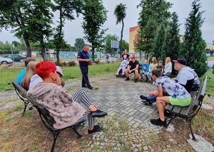 Policjant rozmawia z dziećmi, dzieci siedzą na ławeczkach, policjant stoi miedzy nimi.