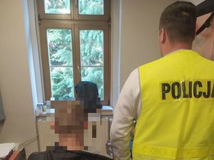 Policjant w kamizelce stoi obok siedzącego mężczyzny w pokoju z oknem