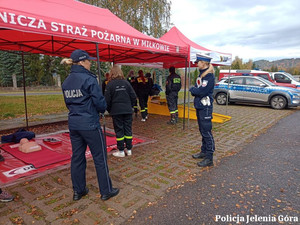 Policjantki stoją przed namiotem, za nimi stojąc strażacy, w tle po prawej stronie policyjny radiowóz