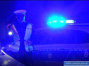 umundurowany policjant w kamizelce odblaskowej i białej czapce oraz z latarką w dłoni stoi na tle oznakowanego radiowozu z włączonymi światłami błyskowymi