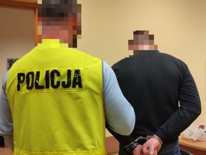 policjant w kamizelce trzyma mężczyznę z kajdankami na rękach trzymanych z tyłu