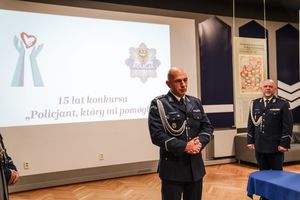 w pomieszczeniu policjant w granatowym mundurze wyjściowym stoi, za nim wyświetlany jest obraz z napisem 15 lat konkursu &quot;Policjant, który mi pomógł&quot;