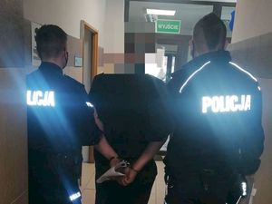 na korytarzy dwóch policjantów w granatowych mundurach z odblaskowymi napisami policja, trzyma stojącą po środku osobę w ciemnej koszulce oraz kajdankach założonych na ręce z tyłu