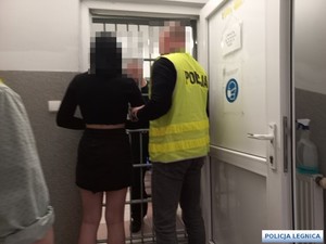 w pomieszczeniu przed kratą mężczyzna w kamizelce odblaskowej z napisem policja trzyma pod rękę kobietę w czarnej spódnicy i czarnej koszulce przed kratami