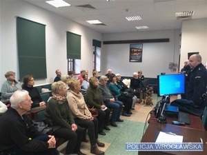 w pomieszczeniu siedzący ludzie i stojący przed nimi policjanci