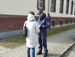 Na dworze na tle budynku policjantka w mundurze wręcza kobiecie ulotkę