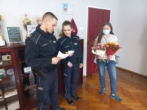Na zdjęciu policjant wraz z policjantką czytają podziękowania. Obok stoi mama dziewczynki wraz z bukietem kwiatów, której córeczka te podziękowania napisała.