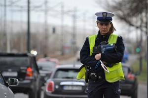 Na tle drogi policjantka w mundurze, białej czapce i kamizelce odblaskowej