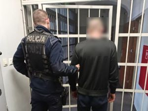 policjant w mundurze trzymający pod rękę innego mężczyznę, w pomieszczeniu przed kratą