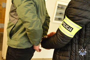 w pomieszczeniu osoba w ciemnej kurtce i odblaskową opaską z napisem Policja na ramieniu, zakłada kajdanki na ręce trzymane z tyłu osobie w zielonej kurtce