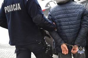 Na dworze policjant w mundurze trzyma pod rękę osobę w ciemnej kurtce kajdankach założonych na ręce z tyłu