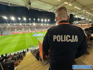 Na zdjęciu policjant stojący tyłem do murawy boiska, na którym rozgrywany jest mecz.