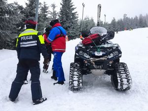 Na zdjęciu skuter narciarski i policjanci pomagający poszkodowanemu.