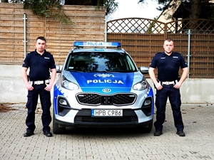 Na zdjęciu dwaj policjanci Mateusz Żukowski oraz Dariusz Kupczyk, między nimi stoi oznakowany radiowóz.