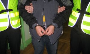 Na zdjęciu dwaj policjanci w kamizelkach odblaskowych prowadzą skutego w kajdanki mężczyznę.