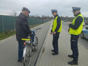 Na zdjęciu dwóch policjantów w kamizelkach odblaskowych poucza starszego mężczyznę stojącego przy rowerze.