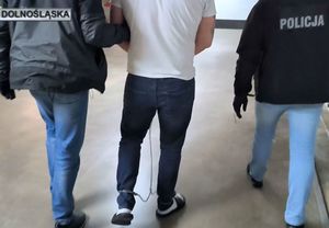 W pomieszczeniu trzy osoby, osoba po prawej w kurtce z napisem &quot;policja&quot;, osoba w środku w białej koszulce z kajdankami założonymi na nogi, osoba z lewej ubrana w ciemną kurtkę.