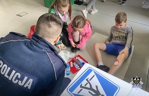 Na zdjęciu widzimy policjanta, który rozmawia z dziećmi o tym jak należy opatrywać rany. Jedno z dzieci ma założony opatrunek na nodze.