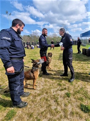 Na zdjęciu policjant zakładający gratulacje i przekazujący puchar jednemu z przewodników psów stojącego z psem. Obok stoi inny przewodnik z psem.