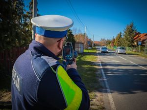 Na zdjęciu policjant stojący przy drodze i mierzący prędkość ręcznym miernikiem prędkości.