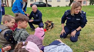 Na zdjęciu policjant i policjantka pokazują dzieciom jak przyjąć bezpieczną pozycję podczas ataku groźnego zwierzęcia. W tle pies policyjny.