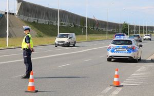 Na zdjęciu policjantka ruchu drogowego stoi na drodze i zatrzymuje pojazdy jadące ulicą. Obok stoi radiowóz.