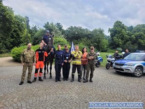 Policjanci i przedstawiciele innych służb stoją na kostce, w tle widać policyjne pojazdy