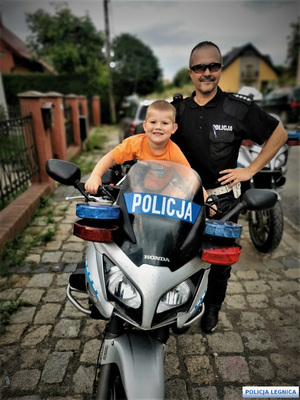 Kilkuletni chłopiec siedzi na motorze policyjnym, obok stoi jego tato - policjant.