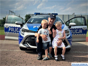 Policjant z malutkimi dziećmi, chłopczykiem i dziewczynką, kucają przy radiowozie.