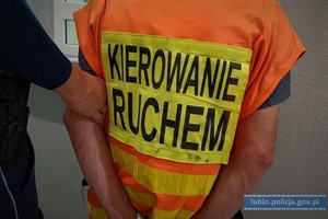 Policjant prowadzi mężczyznę w kamizelce z napisem kierowanie ruchem