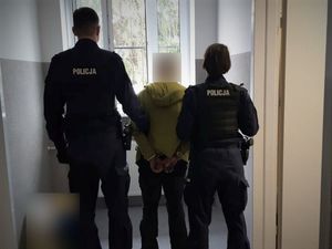 Dwójka umundurowanych policjantów stojąca w pomieszczeniu z oknem z kratami, a pomiędzy nimi stojąca osoba w zielonej bluzie i kajdankach założonych na ręce z tyłu.