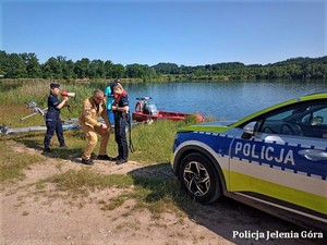 Na pierwszym planie radiowóz policyjny stojący na brzegu jeziora, w tle policjantka mówiąca przez megafon i druga zakładająca kamizelkę ratunkową.