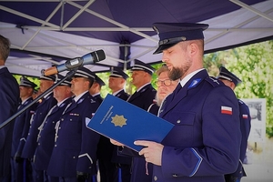 Przemawia policjant prowadzący uroczyste spotkanie, w tle oficjele i zaproszeni goście.