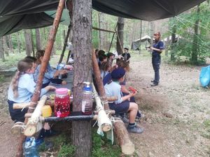 Policjantka mówi do dzieci z obozu harcerskiego i półkolonii siedzących na ławeczce w środku lasu.